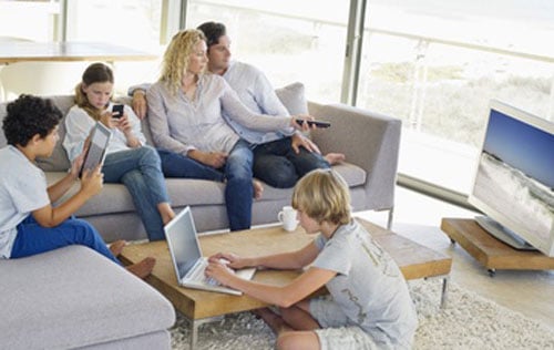 a family sitting in their living room with the parents watching TV,  one son on his laptop, one son on his Ipad and the daughter on her mobile phone 